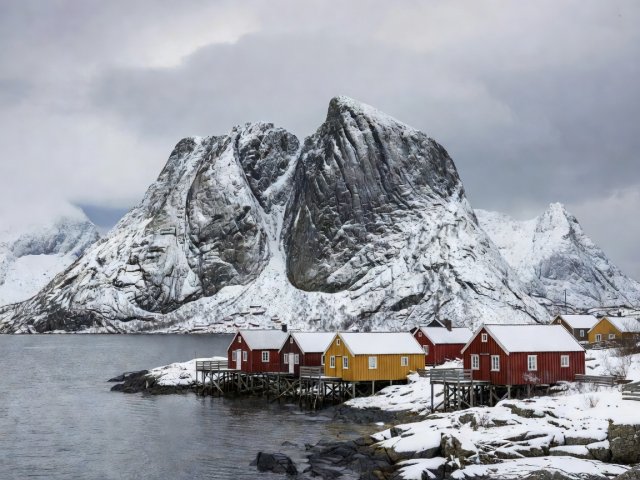 Documentary photograph of jagged snow-dusted granite peaks and traditional rorbuer cabins in Reine, Lofoten Islands, Norway